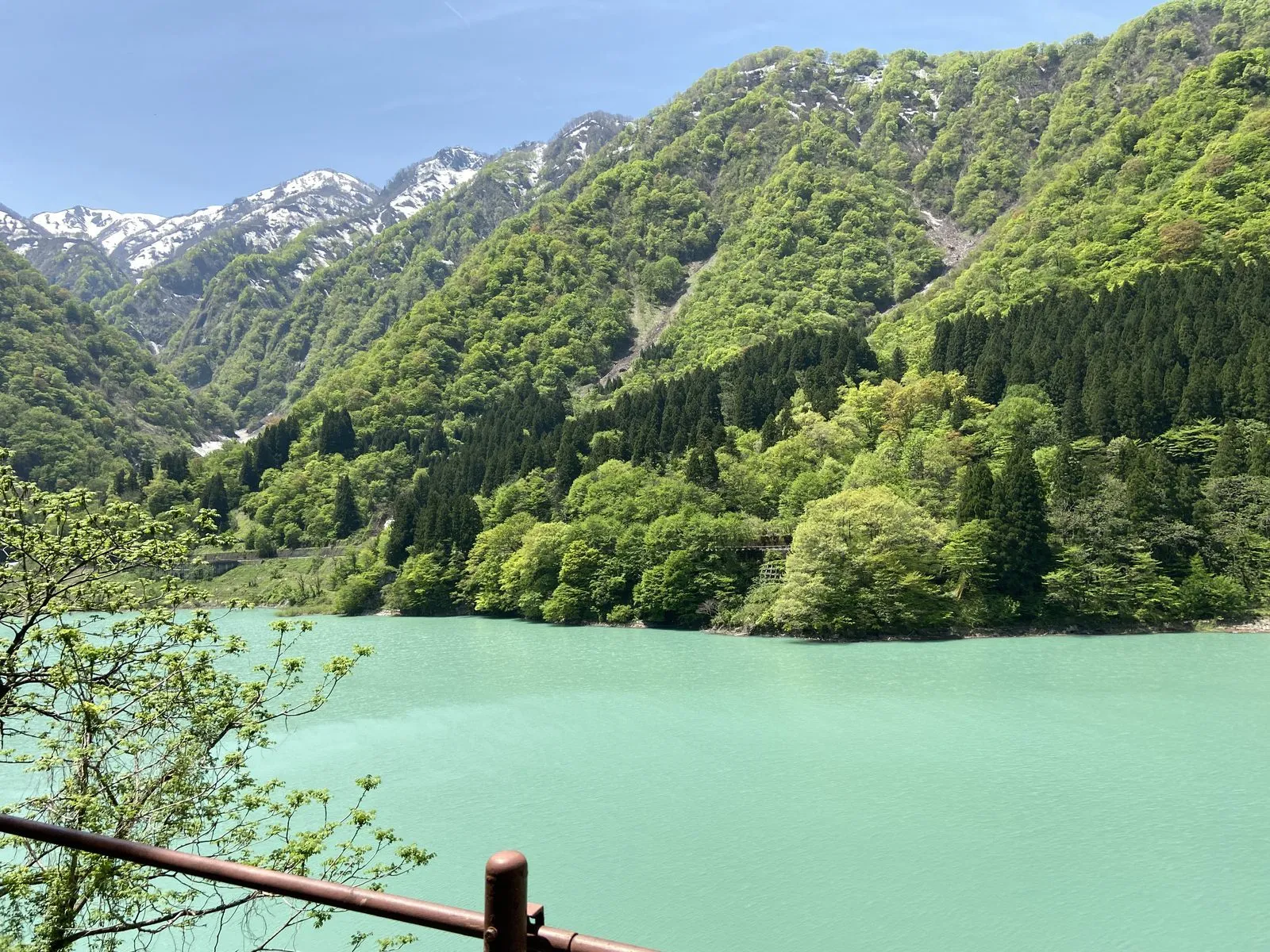 Emerald green dam lake with fresh green mountains and lingering snow, seen from the train